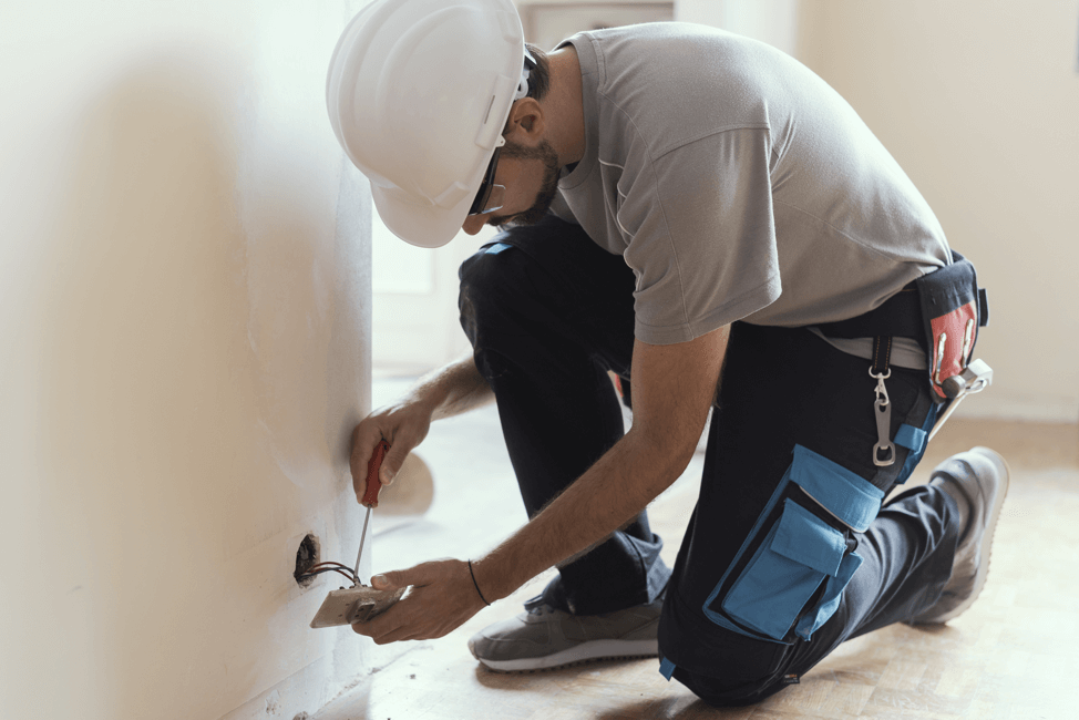 A construction worker in a hard hat kneels while using a screwdriver to repair an electrical outlet on a wall.