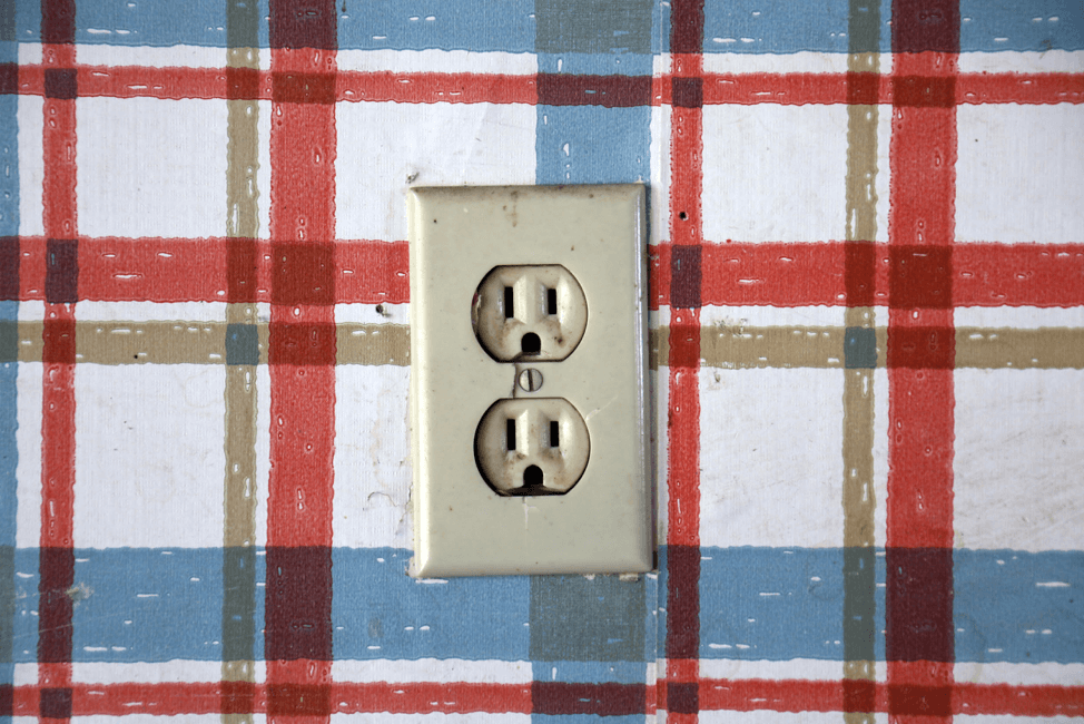 A beige electrical outlet is mounted on a wall with red, blue, and beige plaid wallpaper.