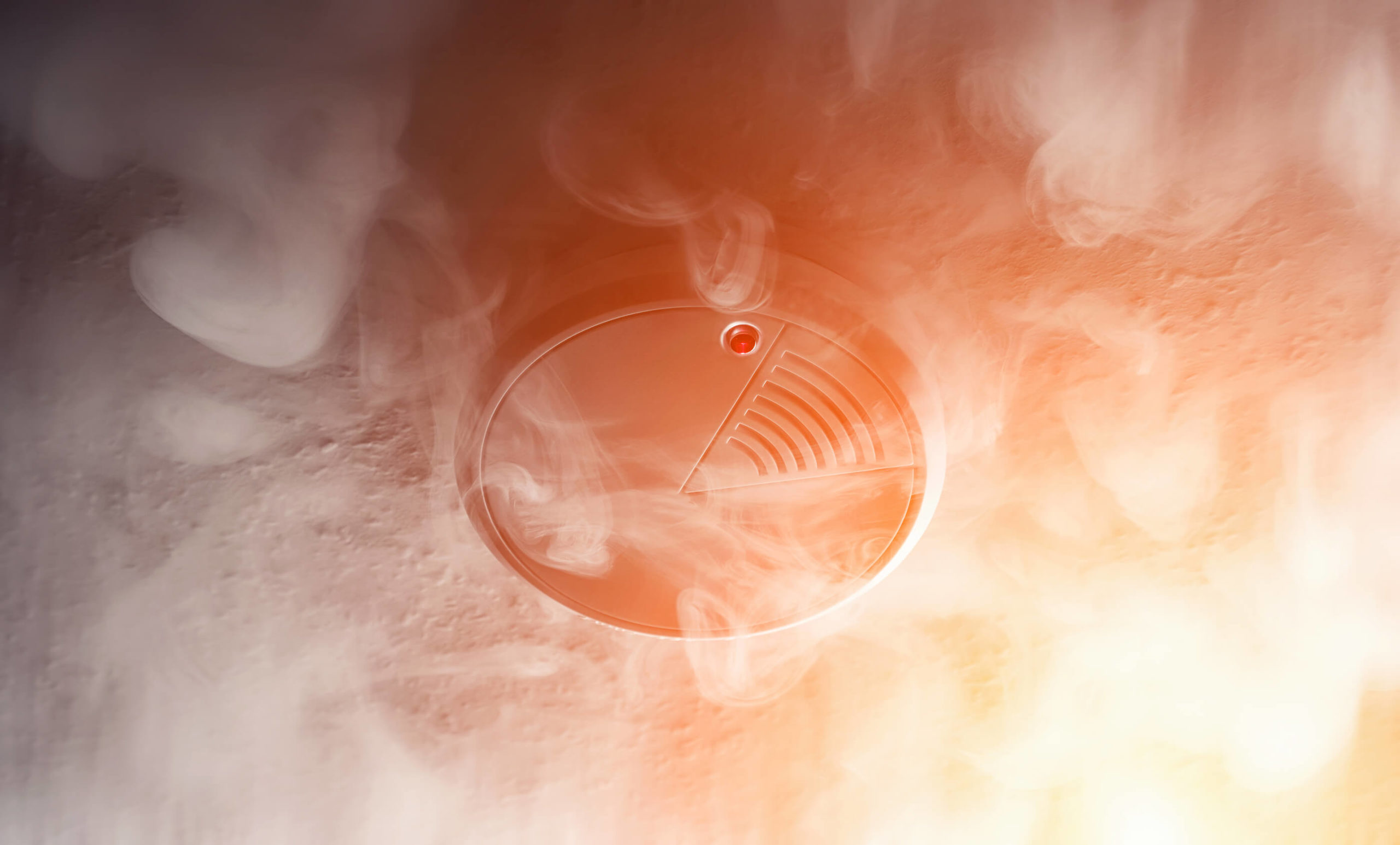 A smoke alarm is mounted on the ceiling surrounded by smoke in a dimly lit environment.