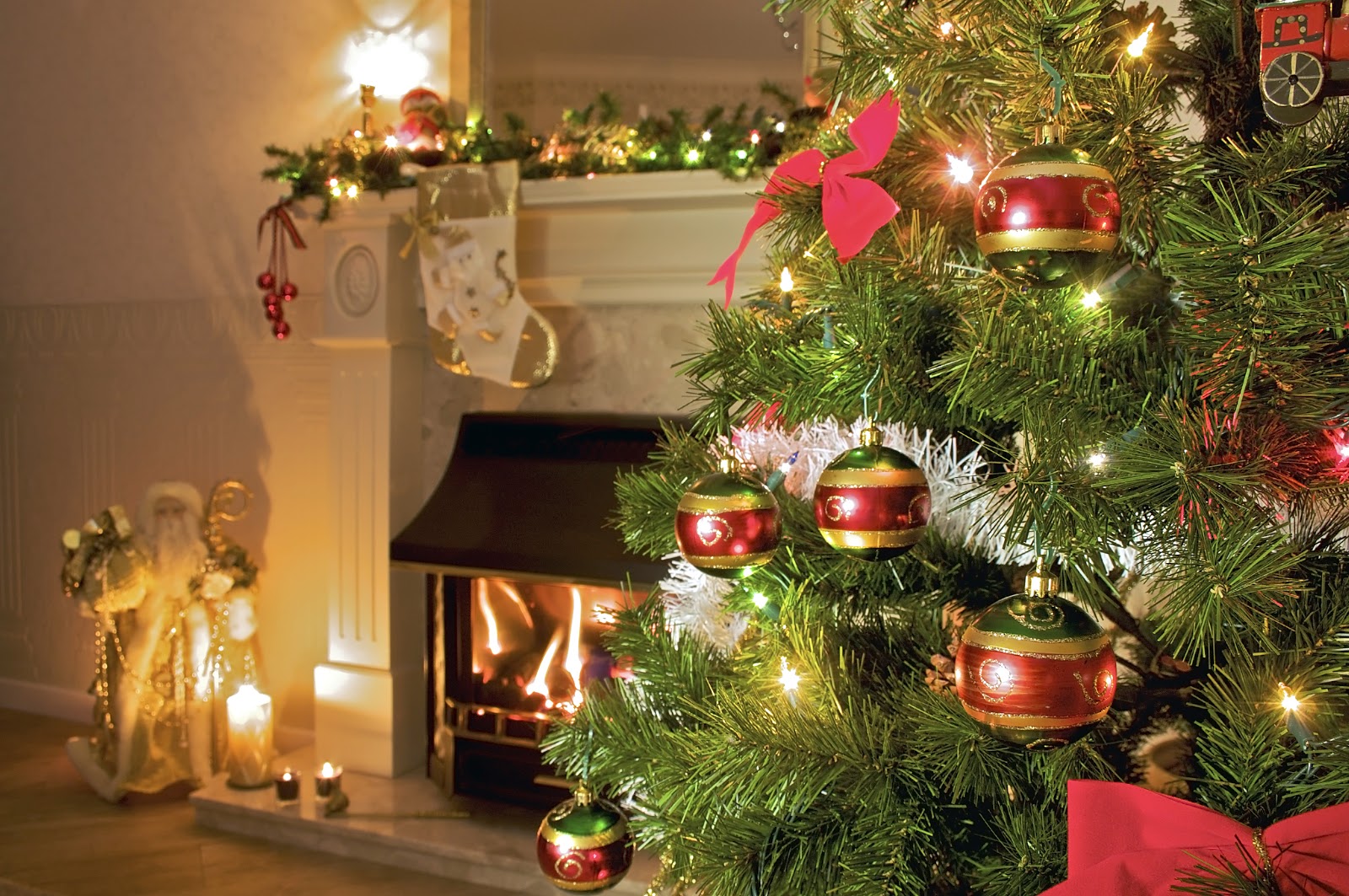 A decorated Christmas tree with red and green ornaments stands beside a lit fireplace and a festive mantle adorned with garlands, candles, and a Santa Claus figure.