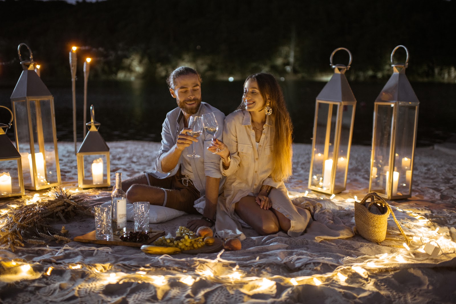 A couple sits on a blanket at a beach picnic at night, surrounded by lanterns and string lights, clinking glasses of wine. A basket, fruit, and a bottle of wine are placed on the blanket.