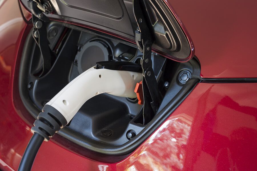 A close-up of an electric car being charged, with a white charging plug connected to the car's charging port. The car is red.