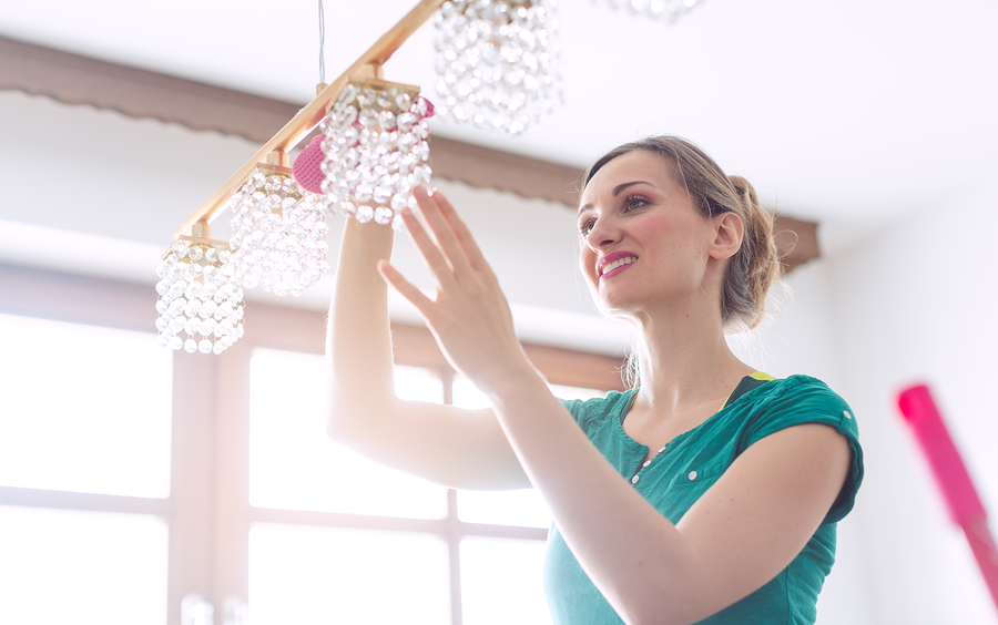 Woman in a green shirt adjusts a crystal light fixture in a bright room with large windows.