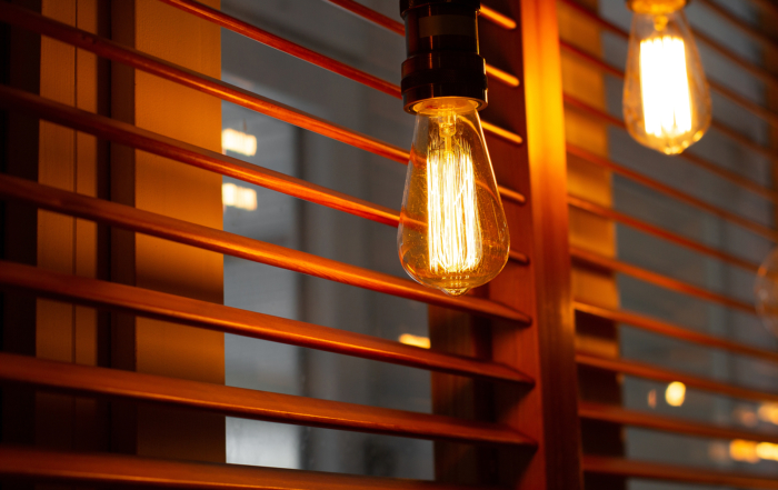 Close-up of a lit vintage-style filament bulb next to wooden window blinds, casting a warm glow.